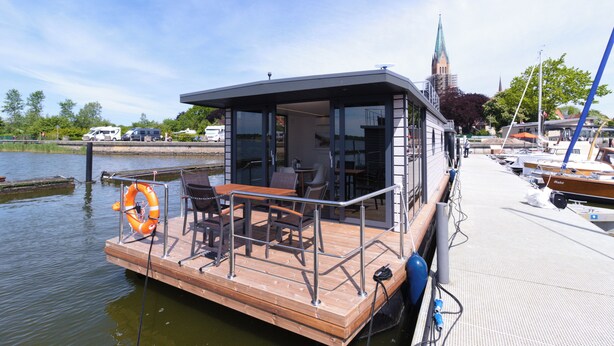 Un restaurante flotante con terraza al aire libre, un flotador salvavidas y vistas al agua, con la aguja de una iglesia al fondo.