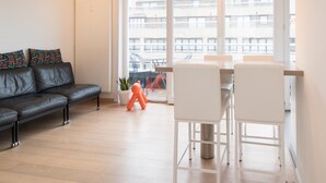 A modern living room with a black leather sofa, white chairs, and a dining table. There is a large window with a view of buildings outside. - Apartment in Ostend Near Beach (Ostend)