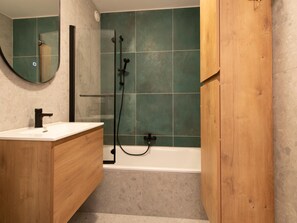 A bathroom with a wooden vanity, a sink, a mirror, a shower with a glass door, and a bathtub. - Apartment in Ostend Near the Beach (Ostend)