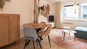 A modern living room with a dining area, featuring a round wooden table, grey chairs, a colorful rug, and a large window with natural light. - Apartment in Ostend Near the Beach (Ostend)