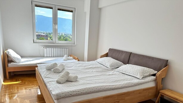 A bedroom with a wooden bed, a window with a view, and a small bedside table. - Air Urban 4-room apartment, balcony, parking, air-conditioned and fully equipped (Sarajevo)