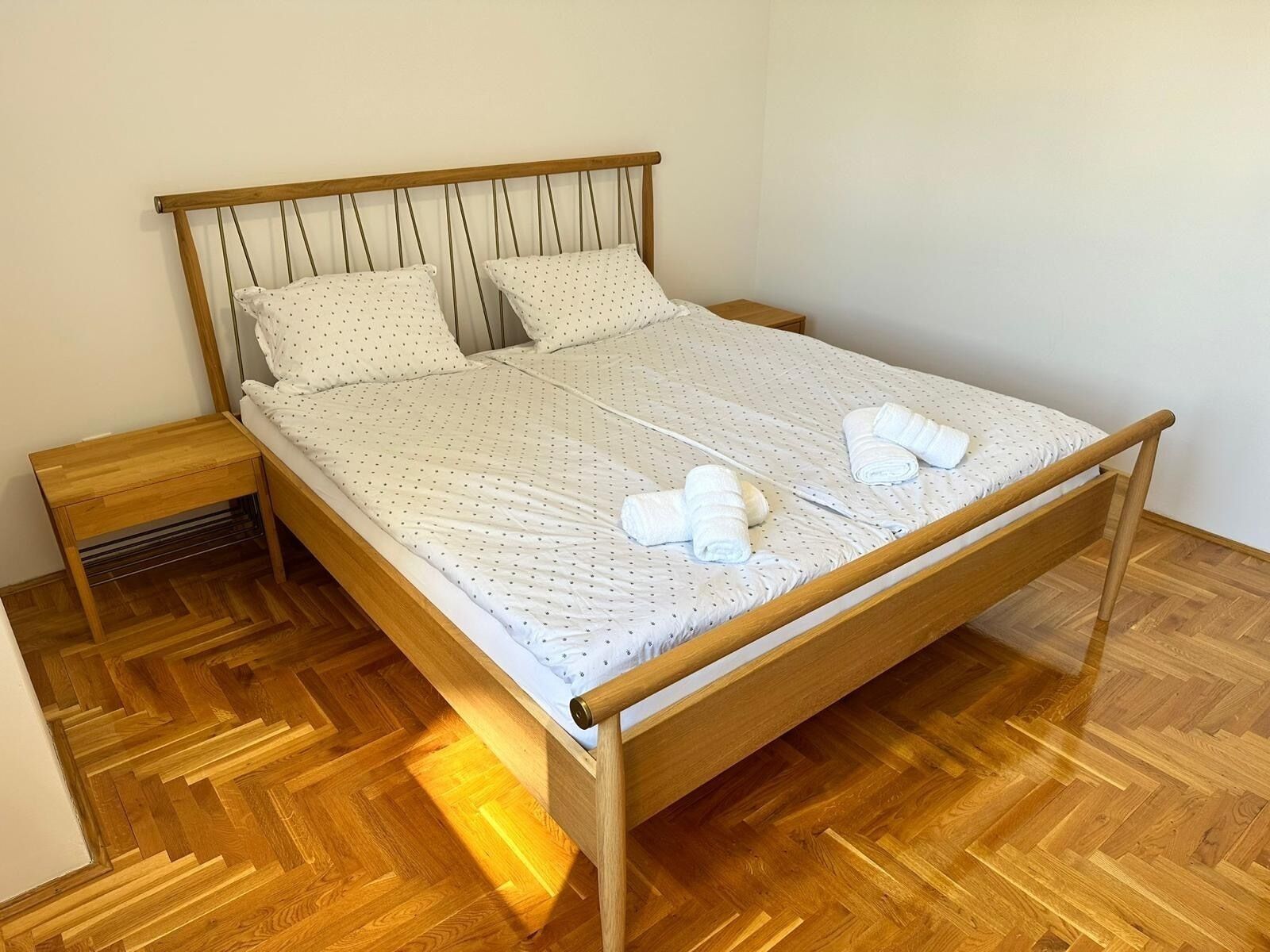 A single bed with a wooden frame and headboard, two pillows, a wooden nightstand, and a wooden floor.