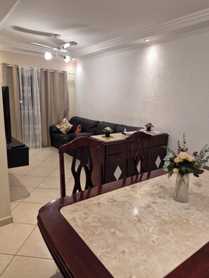 Uma sala de jantar com mesa de mármore, cadeiras de madeira, sofá escuro, ventoinha no teto e televisão fixada na parede.