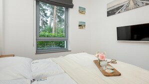 Ein Schlafzimmer mit einem Bett, einem Fenster mit Blick auf Bäume, einem an der Wand montierten Flachbildfernseher und einem kleinen Tisch mit einer Tasse und dekorativen Gegenständen.