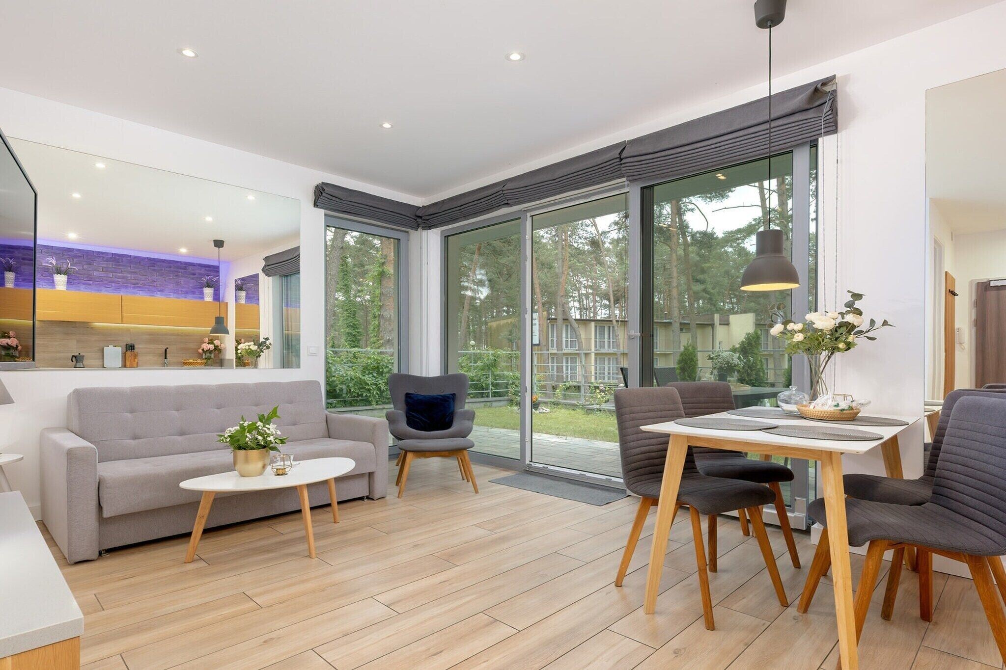 A modern living room with a sofa, a dining table with chairs, and a large window offering a view of the outdoors.