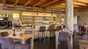 Un restaurant avec des tables et des chaises en bois, un coin bar avec des étagères remplies de verres et de bouteilles, et un plafond haut avec des poutres apparentes.