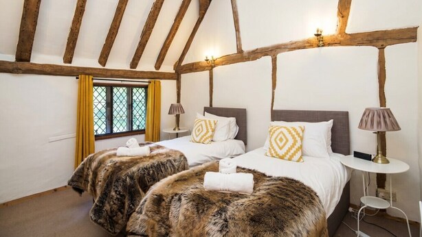 A room with two beds, wooden beams on the ceiling, a window with yellow curtains, and a small table with a lamp. - Spacious East Sussex retreat with tennis court, hot tub, and room for 24 (Whitesmith)