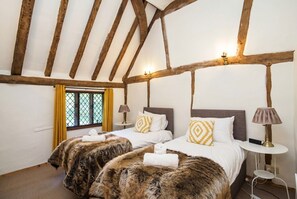 Een kamer met twee bedden, houten balken in het plafond, een raam met gele gordijnen, en een kleine tafel met een lamp.