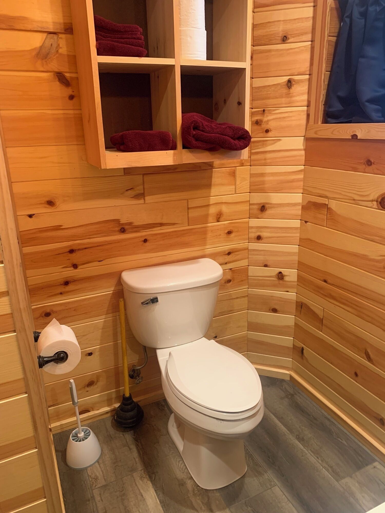 Un baño con paneles de madera, un inodoro, un estante con toallas y un dispensador de papel higiénico.