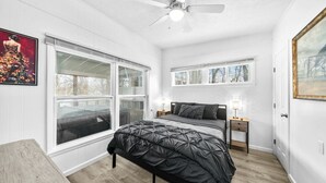 Un dormitorio con cama, dos mesitas de noche, ventilador de techo y ventanas amplias.
