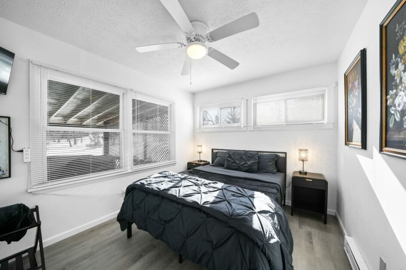 A bedroom with a bed, bedside tables, a ceiling fan, and large windows with blinds.