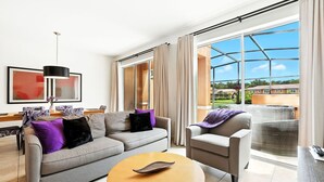 Ein modernes Wohnzimmer mit grauem Sofa, rundem Couchtisch und grauem Sessel. Es gibt ein großes Fenster mit Vorhängen, eine schwarze Pendelleuchte und ein gerahmtes Bild an der Wand.