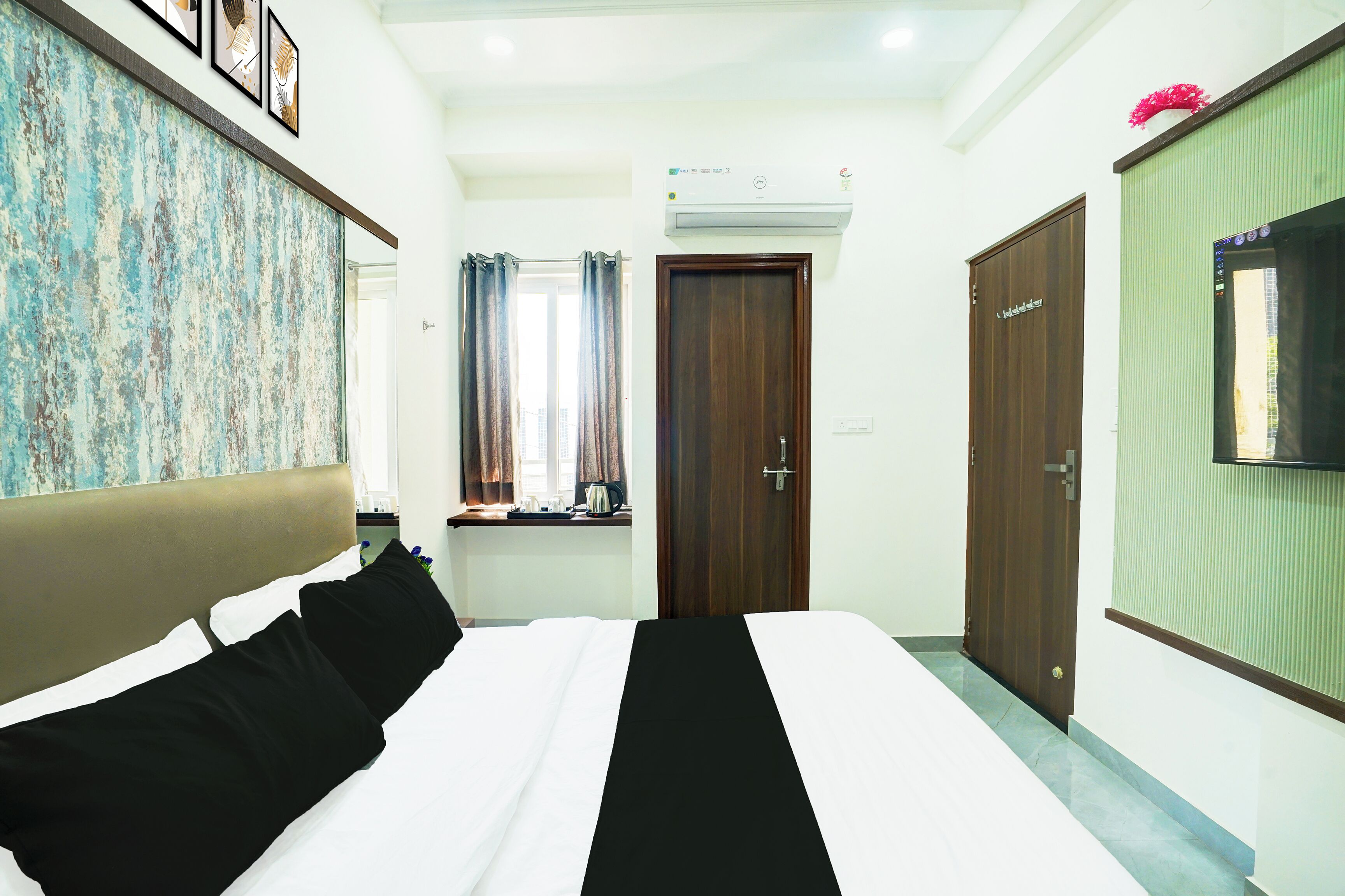 A hotel room with a bed, a window with curtains, a wooden door, and a wall-mounted TV.