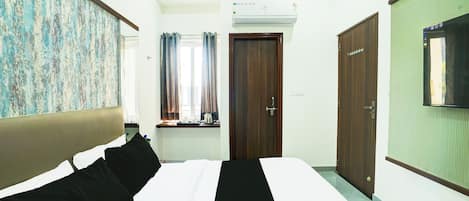 A hotel room with a bed, a window with curtains, a wooden door, and a wall-mounted TV.