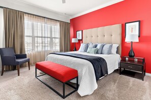 Habitación de hotel con una cama, una silla, un banco, una mesita de noche, una lámpara y una ventana con cortinas.