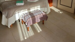 A neatly made bed with pillows and a blanket, a bedside table with a lamp, a small wooden stool, and a painting on the wall.