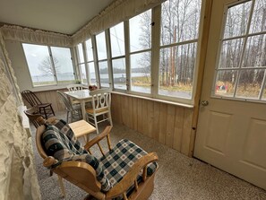 Une pièce chaleureuse au décor intérieur en bois, avec une table à manger et des chaises, un banc rembourré et de grandes fenêtres offrant une vue sur les arbres et un plan d’eau.