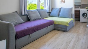 Un espace de vie moderne et compact, comprenant un canapé-lit, un lave-linge et une grande fenêtre avec des rideaux.