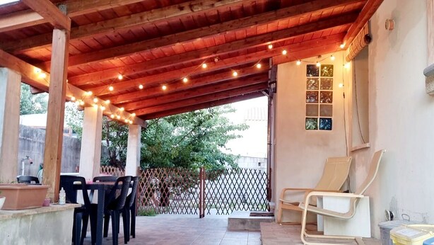 Une terrasse couverte, illuminée de guirlandes lumineuses, avec une table et des chaises, un fauteuil de relaxation et une fenêtre à carreaux.