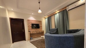 Uma sala de estar moderna com uma TV de tela plana fixada em uma parede de madeira, um sofá azul, uma porta de madeira escura e piso ladrilhado.