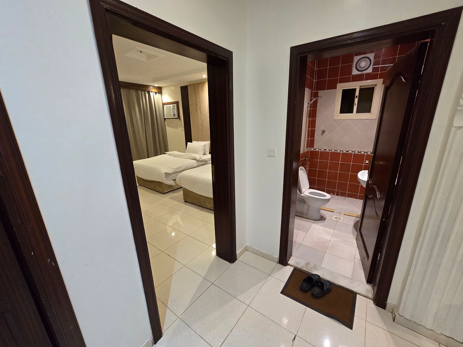 A hotel room with a bed, a bathroom with a toilet and sink, and a hallway with a pair of shoes on a mat.
