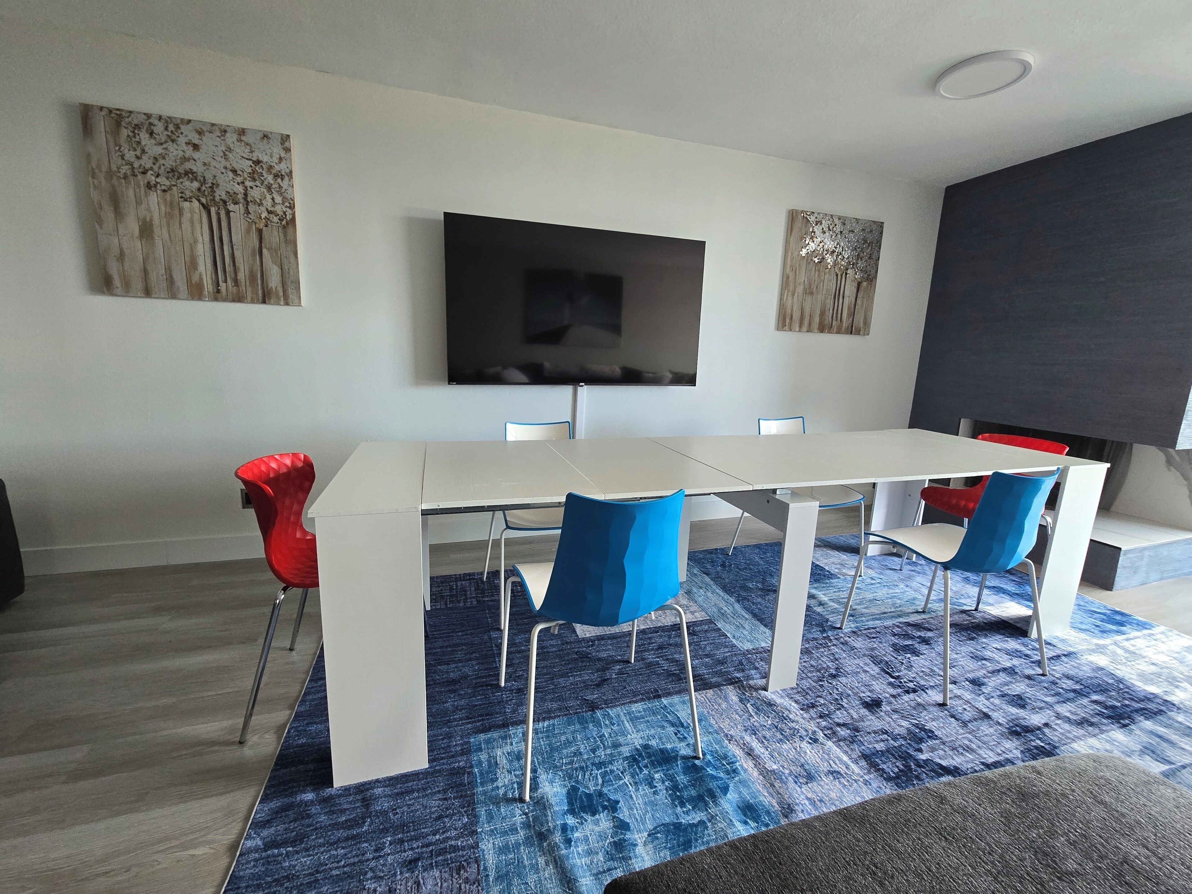 Uma área de refeições moderna com mesa branca e cadeiras azuis, uma televisão de tela plana na parede e obras de arte acima da mesa.