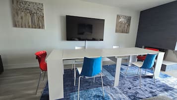 Uma área de refeições moderna com mesa branca e cadeiras azuis, uma televisão de tela plana na parede e obras de arte acima da mesa.