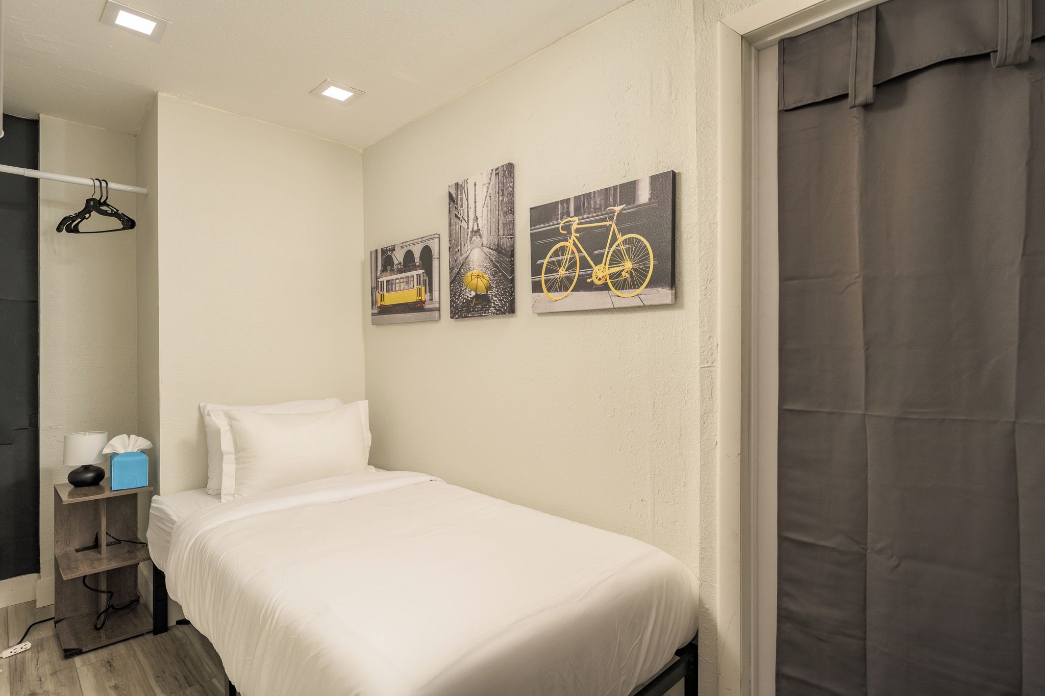 Quarto de hotel com uma cama de solteiro, mesa de cabeceira com abajur e lenços, e três quadros na parede.