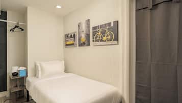 Quarto de hotel com uma cama de solteiro, mesa de cabeceira com abajur e lenços, e três quadros na parede.