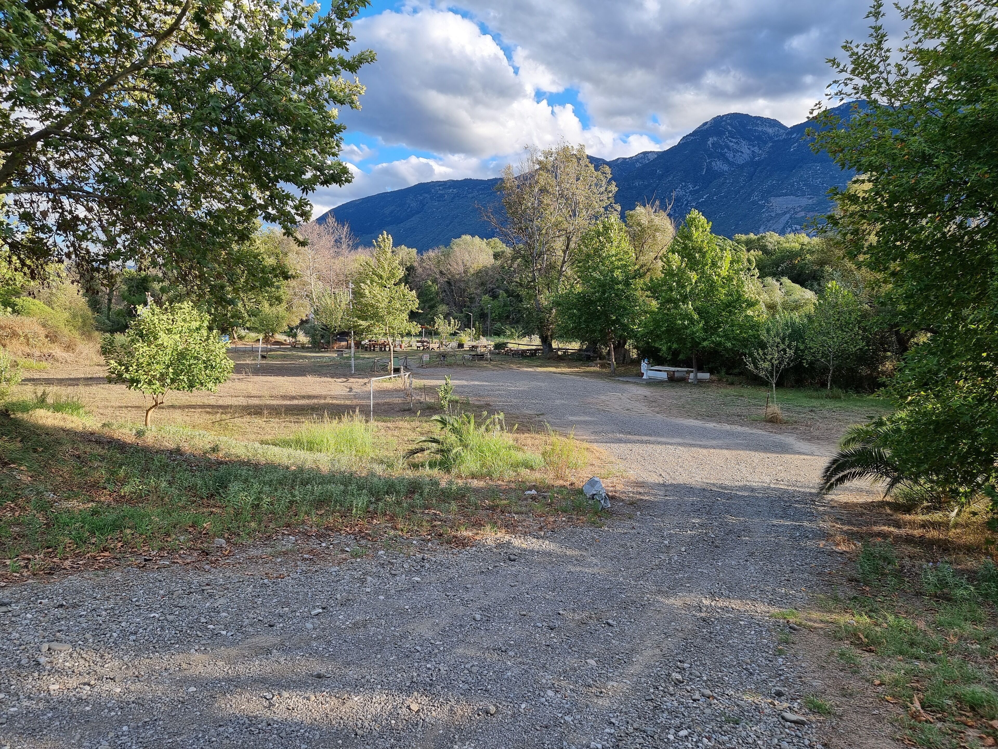 A gravel path leading to a picnic area with tables and benches, surrounded by trees and mountains in the background.