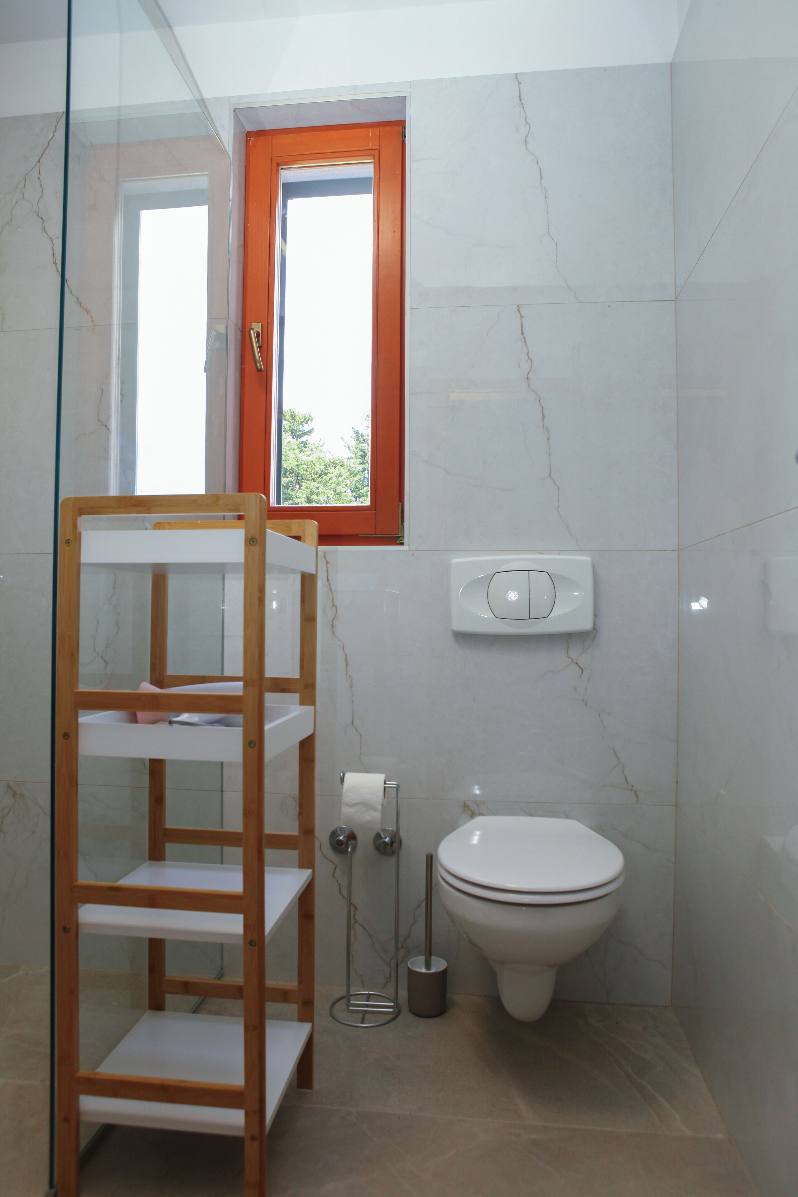 A bathroom with a wooden shelf, a toilet, a bidet, and a window with an orange frame.