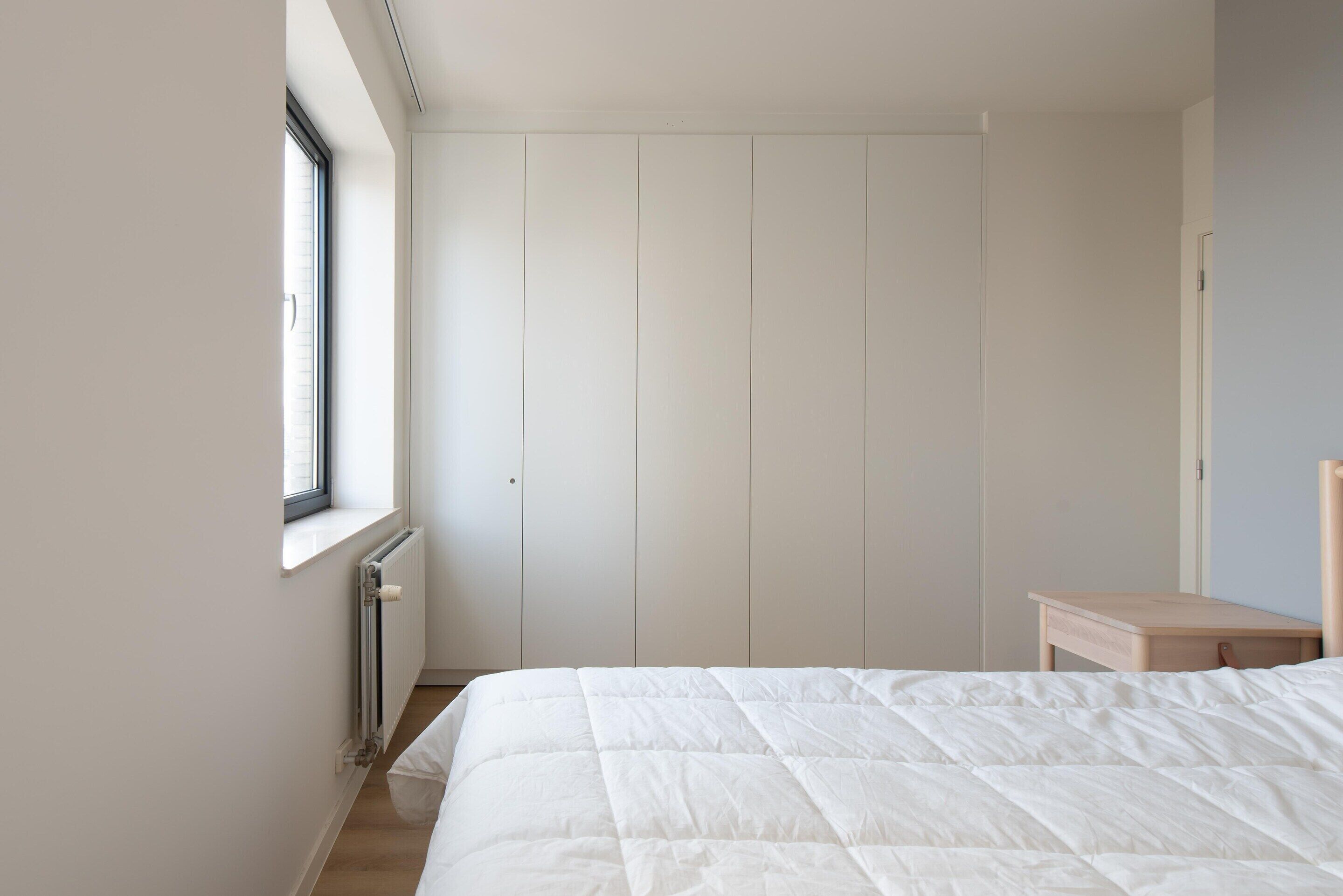 A bedroom with a large bed, a window, a radiator, and a wooden nightstand.
