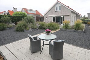 Un patio con mobiliario de mimbre y una mesa, rodeado de un jardín con diversas plantas y un camino de grava.