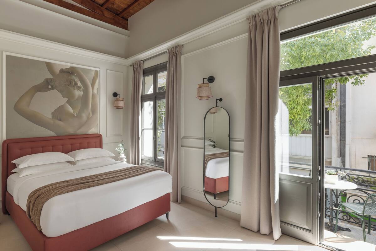 A bedroom with a large bed, a wall-mounted mirror, and a view of the outside.
