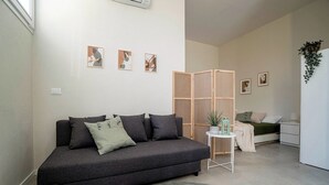A modern living room with a grey sofa, a small round rug, and a bed with a green blanket. There are abstract paintings on the wall and a plant on the sideboard. - Casa del Falegname · Abete Loft with kitchen · 3 guests (Gaiana di Castel San Pietro Terme)