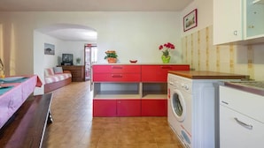 A compact living space with a sofa, a washing machine, and a small kitchen area with red cabinets and a countertop.