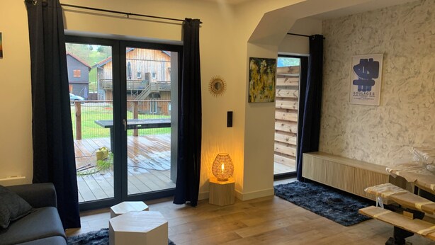 A modern living room with a sofa, a coffee table, and a dining area with benches. There is a sliding glass door leading to a patio with a wooden deck and a view of a residential area. - Residence "La Menuiserie" 8 guests, spa sauna (2) (GÉRARDMER)