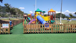 Un parque infantil con valla de madera, un letrero que indica restricciones de altura y una playa al fondo.