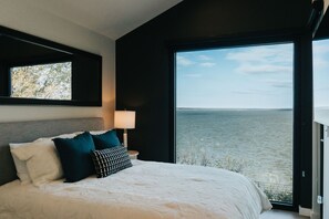 Camera da letto con un letto grande, un comodino, una lampada e vista sull'acqua tramite un'ampia finestra.