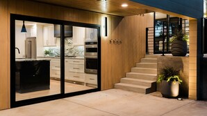Une cuisine moderne dotée d’un grand îlot central, d’appareils électroménagers intégrés et d’une porte-fenêtre coulissante donnant sur un espace extérieur agrémenté d’un escalier et de plantes en pot.
