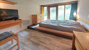 Quarto com uma cama, televisão, escrivaninha de madeira e janela com cortinas.