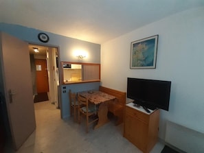 Pieni huone, jossa on pieni keittiö, ruokapöytä tuoleineen, televisio puukehikossa ja seinällä kehystetty taulu.