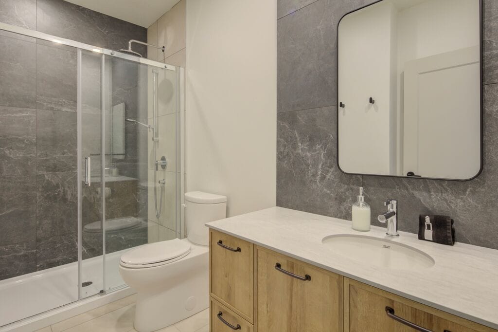 Un bagno moderno con doccia vetrata, un water bianco, parete rivestita in marmo, uno specchio grande e un mobile bagno in legno con lavabo.