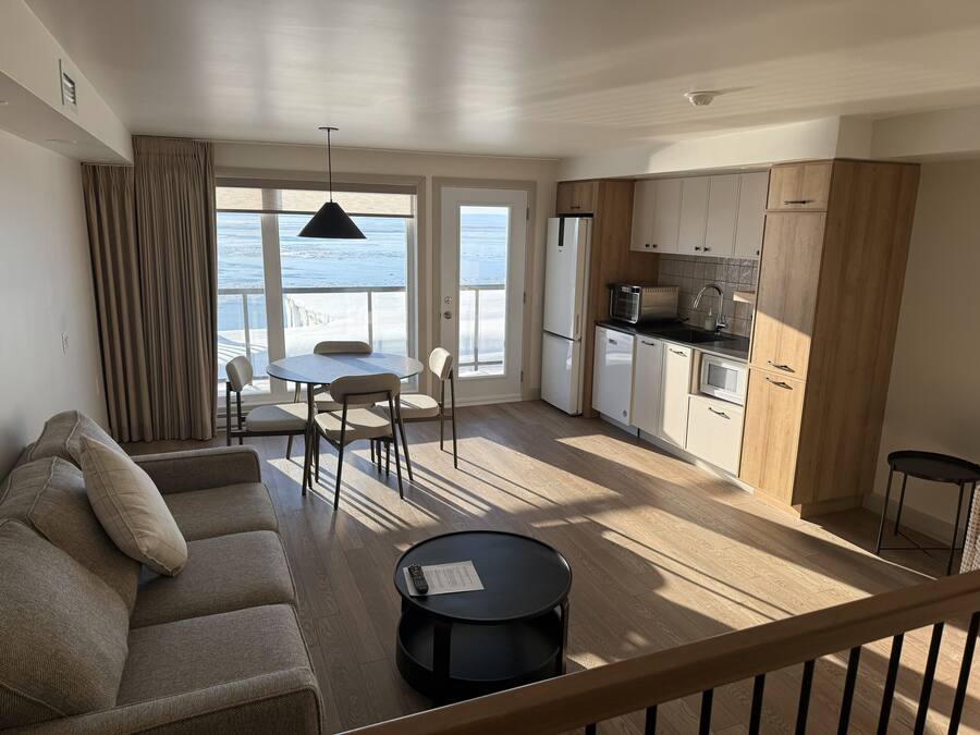 A modern living room with a sofa, a dining table, and chairs, a kitchen with wooden cabinets, a refrigerator, and a microwave.