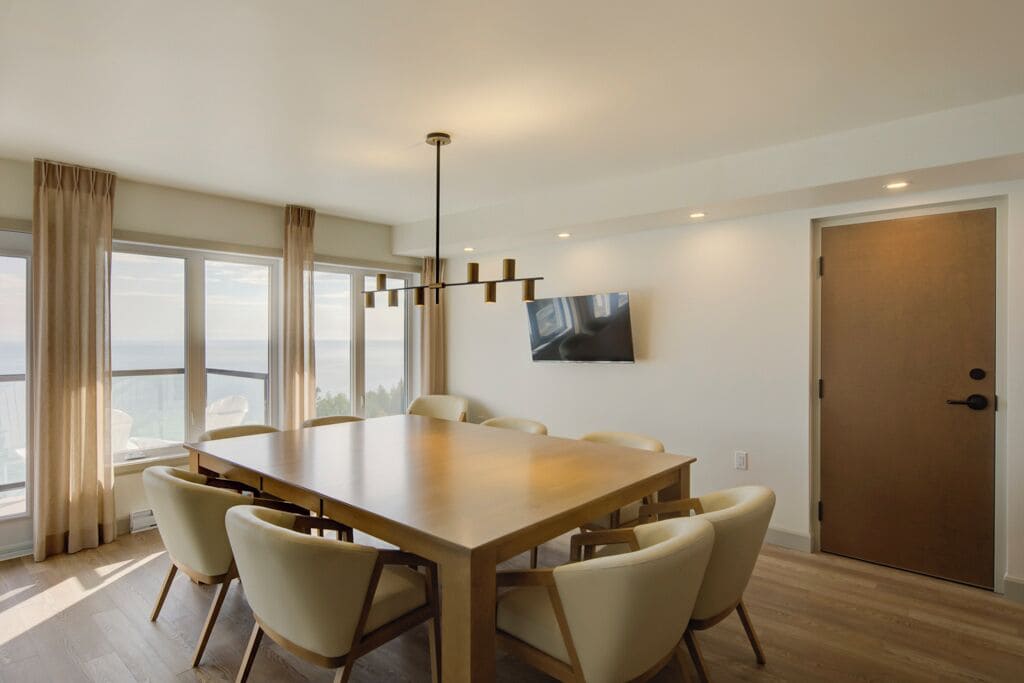 A modern dining room with a large wooden table and six chairs, large windows with curtains, a mounted TV, and a wooden door.
