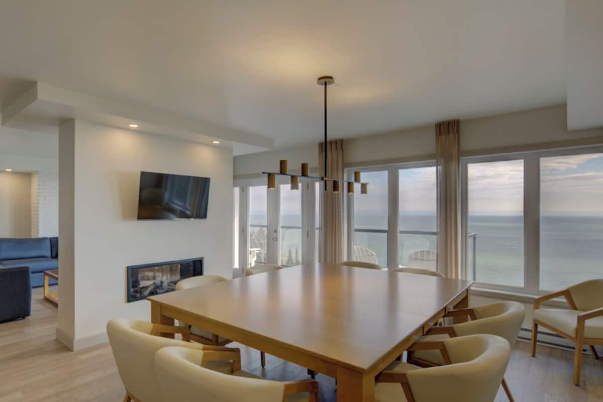 Een ruime eethoek met een grote houten tafel en stoelen, een aan de muur bevestigde televisie, en grote ramen met uitzicht op de zee.