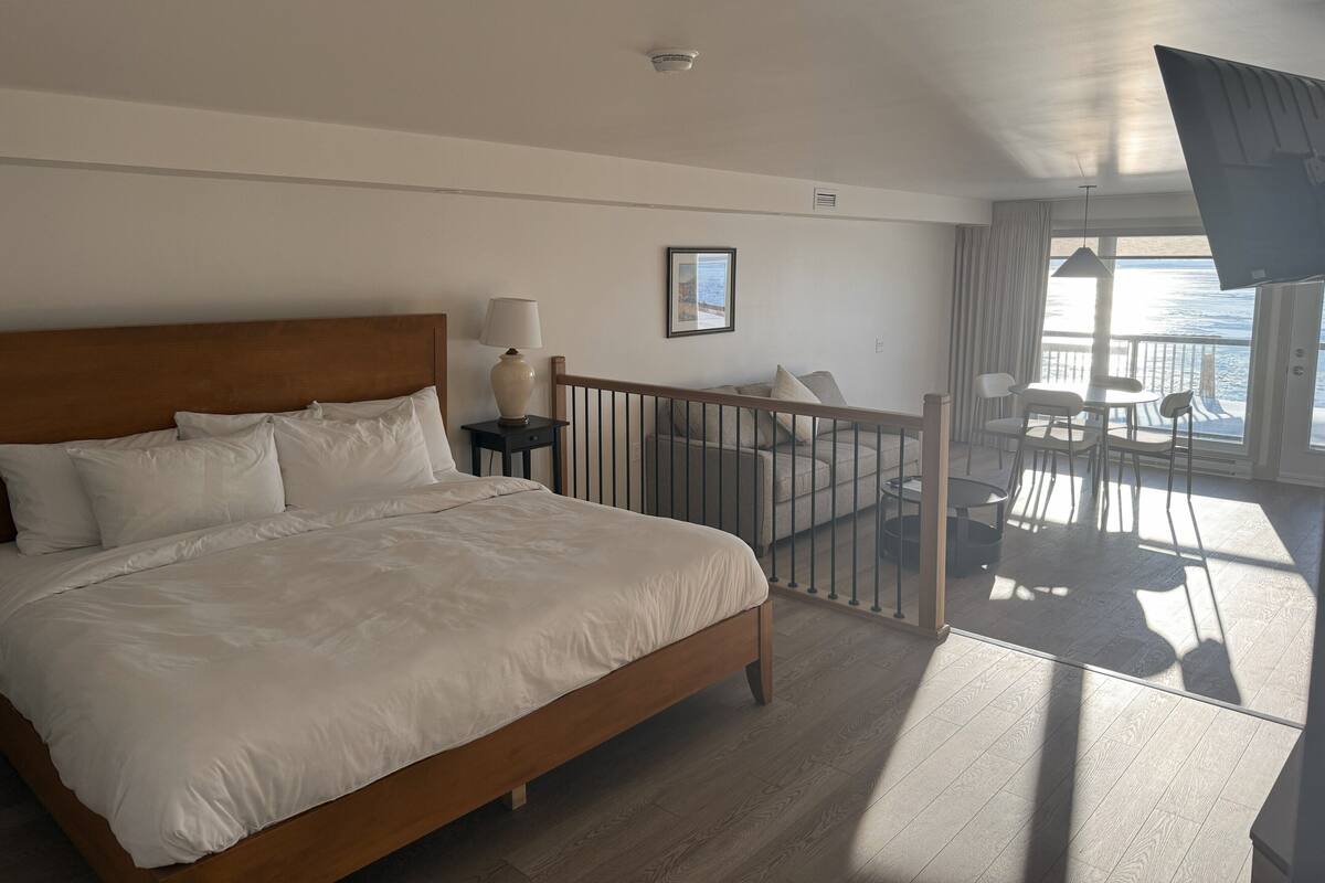 Een hotelkamer met een groot bed, een nachtkastje, een lamp, een lijst met een afbeelding, een balkon met uitzicht op zee en een plafondventilator.