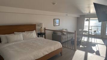 A hotel room with a large bed, a nightstand, a lamp, a framed picture, a balcony with a view of the sea, and a ceiling fan.