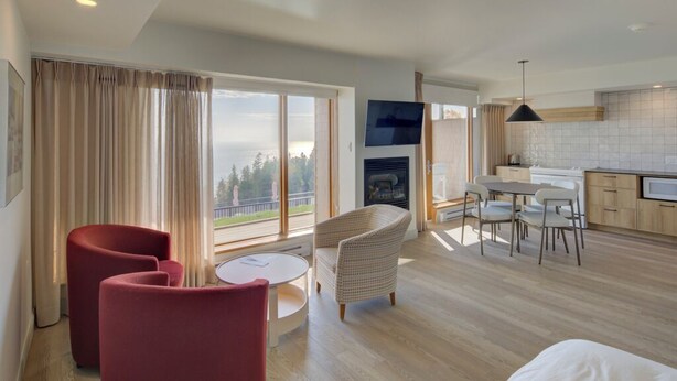 A modern hotel room with a large window, a fireplace, a dining area, and a kitchen. - La Pinsonnière (La Malbaie)