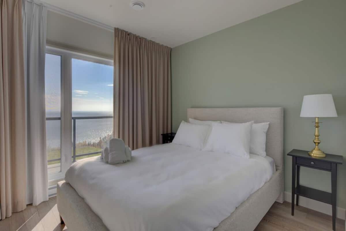 Een groot bed met witte lakens, een nachtkastje met een lamp, en uitzicht op zee door een raam.